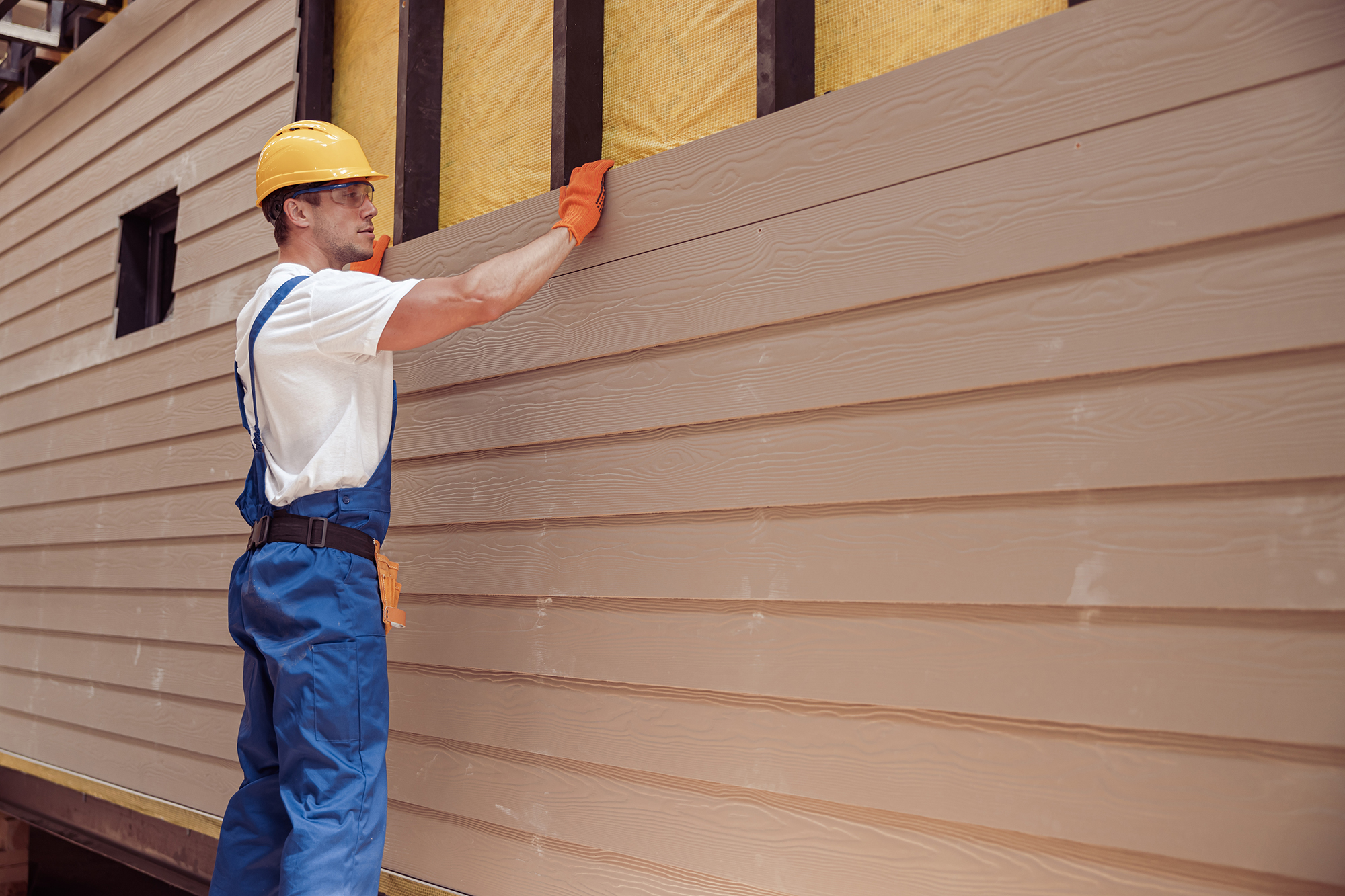 A construction worker in a yellow hard hat and blue overalls guides a long piece of light brown siding into place on a building, illustrating a step in Spearfish Siding Installation by Wegner Roofing & Solar.
