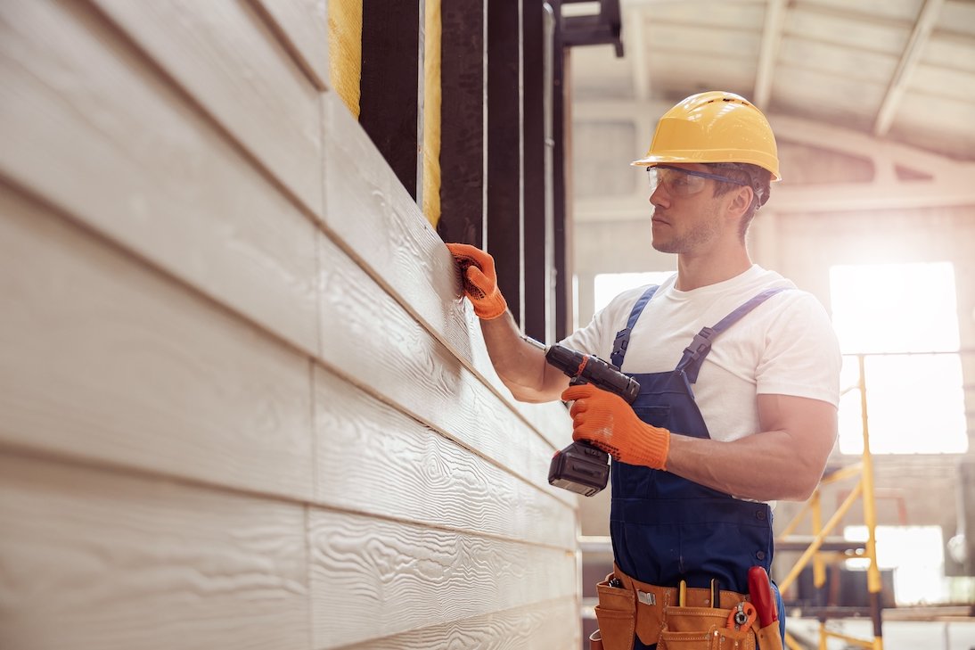 A construction worker in a yellow hard hat and orange gloves uses a power drill to install light-colored horizontal vinyl siding on a building, demonstrating Spearfish Siding Installation by Wegner Roofing & Solar.