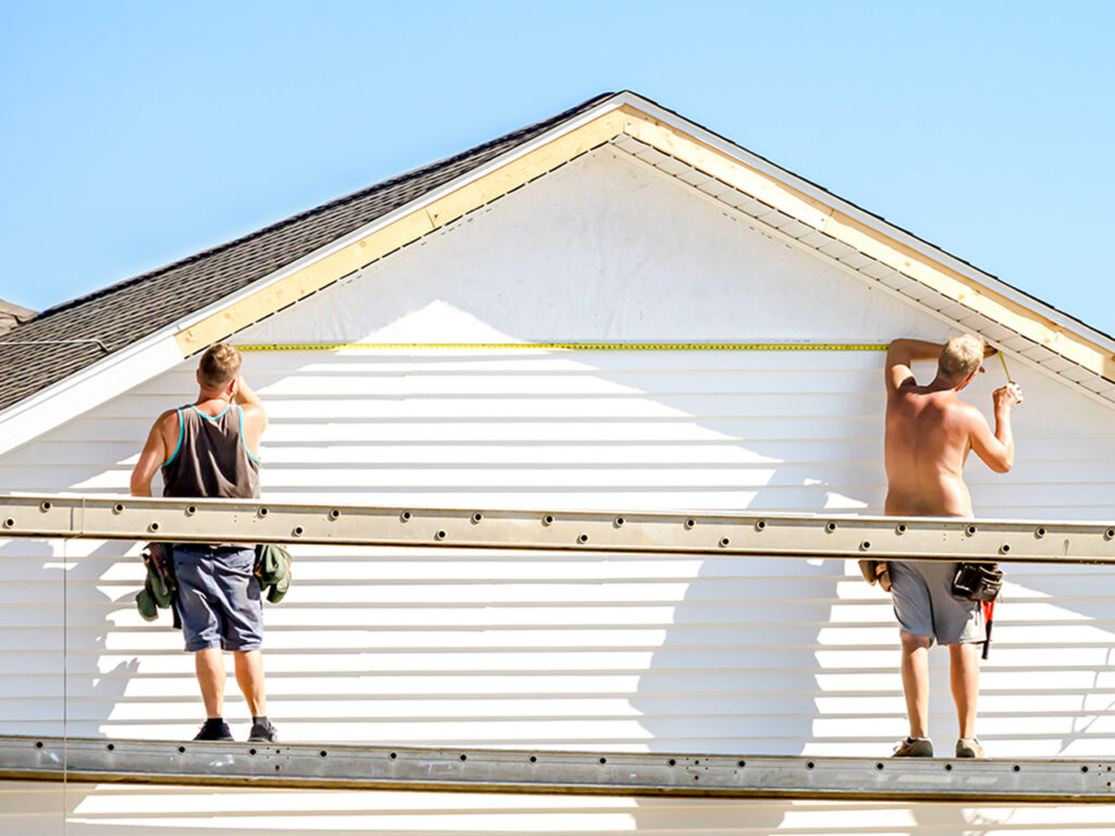 Two professional Spearfish Siding Installers from Wegner Roofing & Solar are standing on a scaffolding, installing white vinyl siding on the gable end of a house. One installer is holding a measuring tape across the upper section of the completed siding, while the other marks a piece of trim near the roof line on a clear, sunny day.