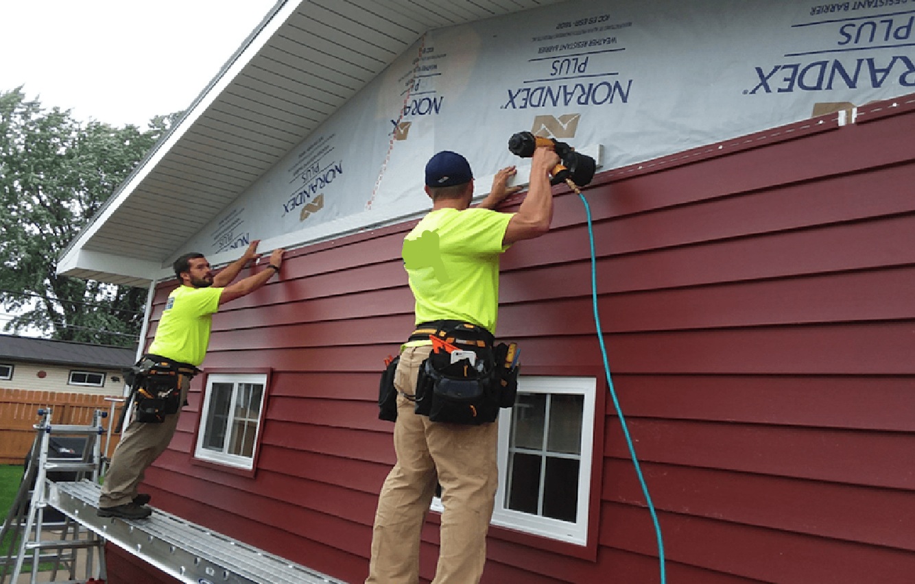 Two Spearfish Vinyl Siding Installers from Wegner Roofing & Solar working on a two-story home with red vinyl siding. The installer on the right is using a pneumatic nailer to secure the top row of red vinyl siding below the gable end, which is currently covered with a Norandex house wrap. The second installer is standing on a ladder to the left, aligning a piece of siding.