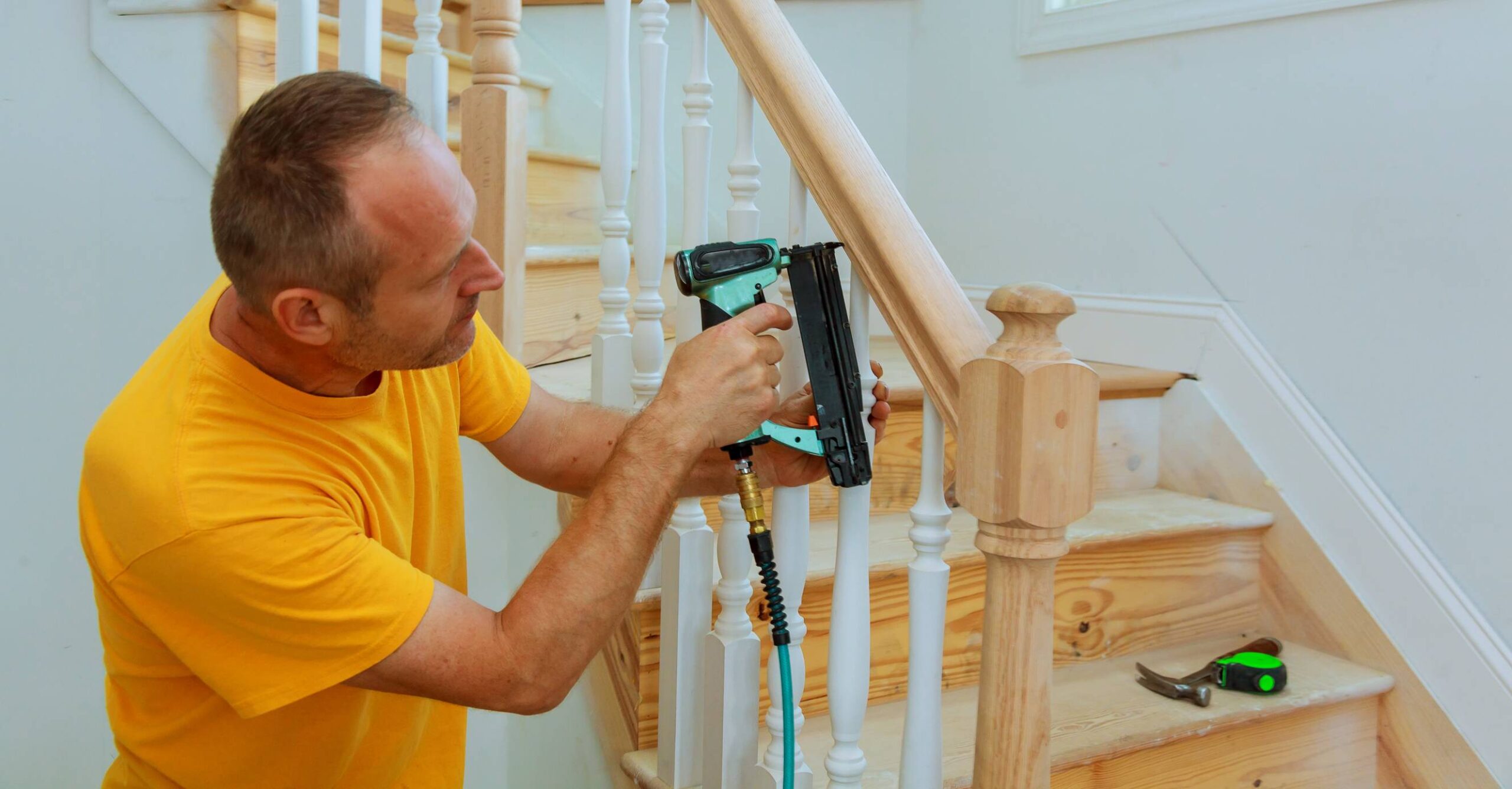 A craftsman in a yellow t-shirt is using a pneumatic nail gun to fasten a wooden handrail to the newel post of a staircase. The staircase has wooden treads and white balusters, indicating ongoing finish carpentry work. Tools like a hammer and tape measure are visible on the stair tread. This image illustrates Staircase and Handrails Installation Roswell GA by Peachy Flooring Solutions.
