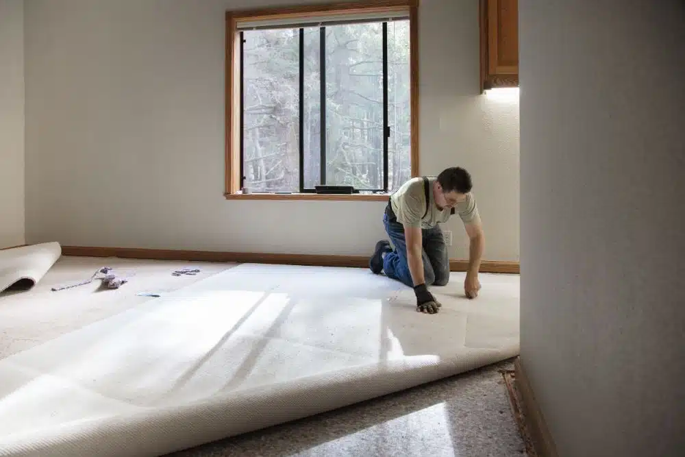 Installer on his knees unrolling new light-colored carpet in a room with a large window during North Atlanta GA Carpet Installation.